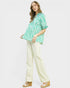 Woman wearing a green patterned top and white pants on a white background