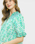 Woman wearing a green and white patterned blouse with ruffled sleeves on a plain background