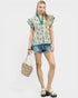 Woman wearing a floral blouse and denim shorts, holding a woven bag on a white background