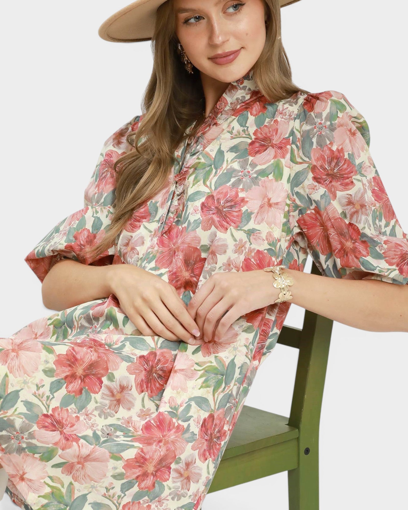 A woman wearing a floral midi dress with puff sleeves and a high collar, seated on a green wooden chair against a light gray background.