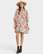 A woman modeling a long-sleeve floral shirt dress with a high collar, paired with a wide-brimmed straw hat and brown ankle boots, against a light gray studio background.