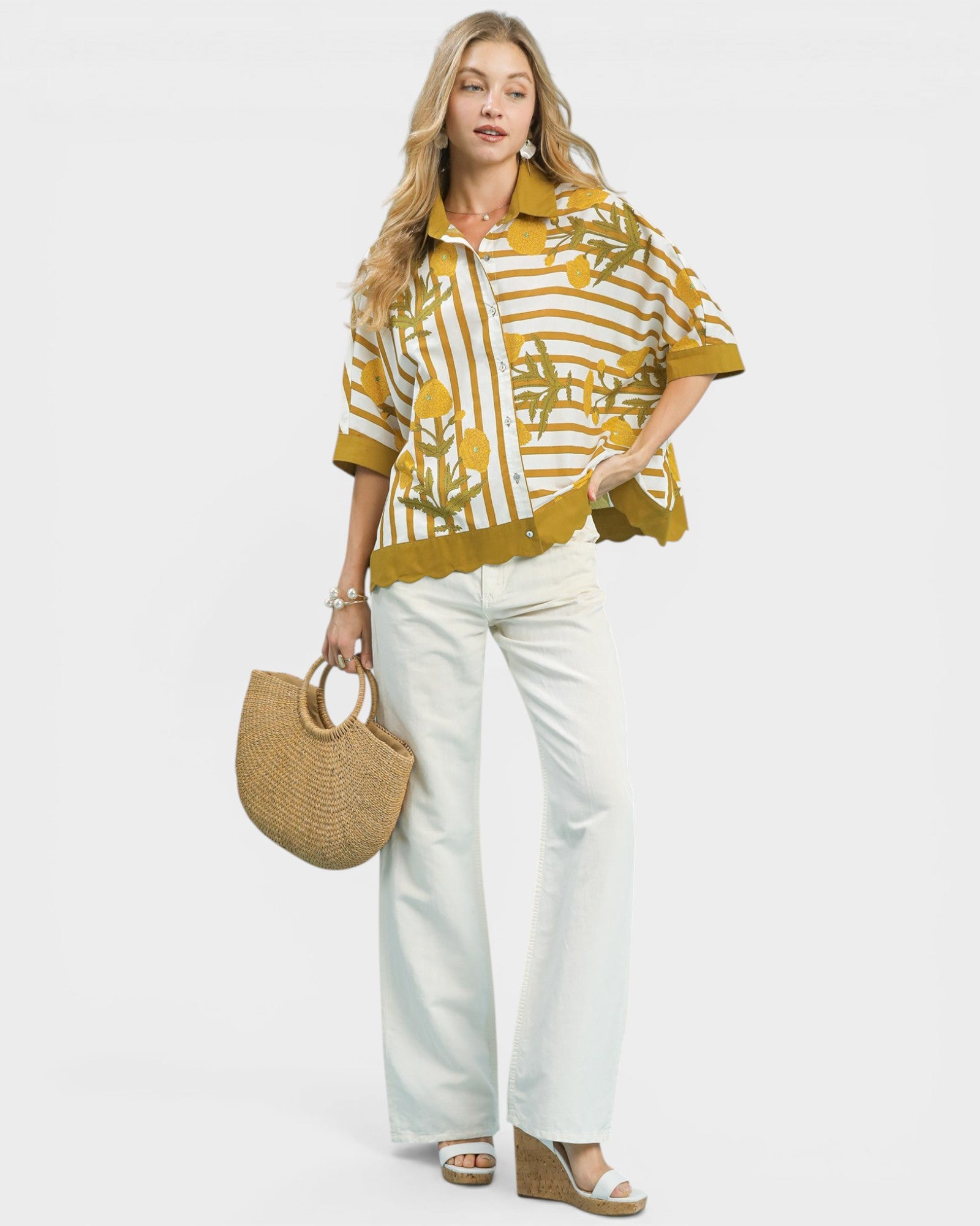 Woman wearing a mustard yellow and white patterned shirt with white pants, holding a straw bag on a white background.