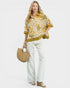 Woman wearing a mustard yellow and white patterned shirt with white pants, holding a straw bag on a white background.