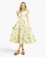 A woman modeling a tiered midi dress with a light cream base and a delicate multicolor floral print, featuring a light blue button-up collar and ruffled sleeves, paired with brown platform sandals against a light gray background.