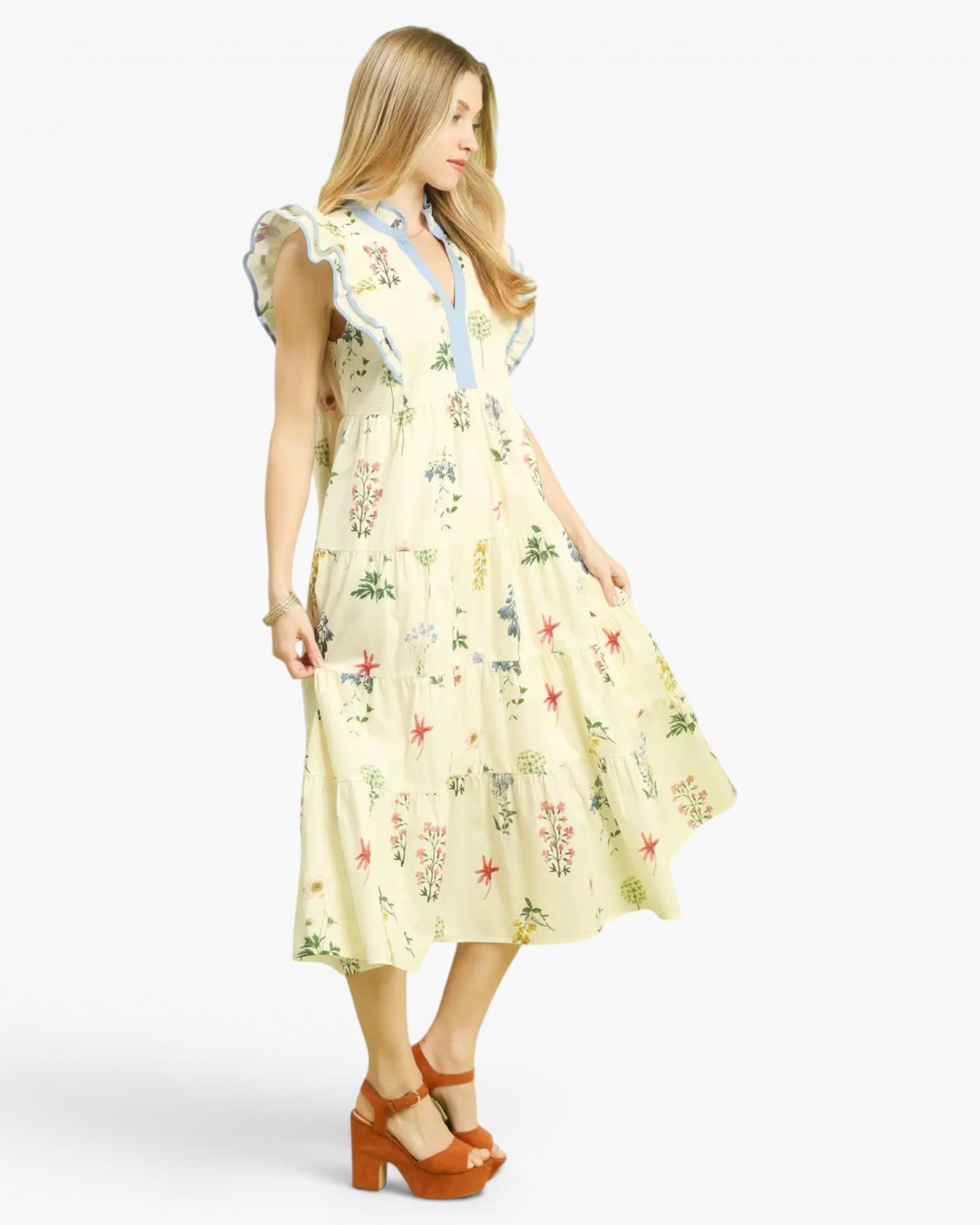 A woman modeling a light yellow midi dress with a colorful floral and starburst print, featuring ruffled cap sleeves and a light blue collar, paired with brown platform sandals against a plain white background.