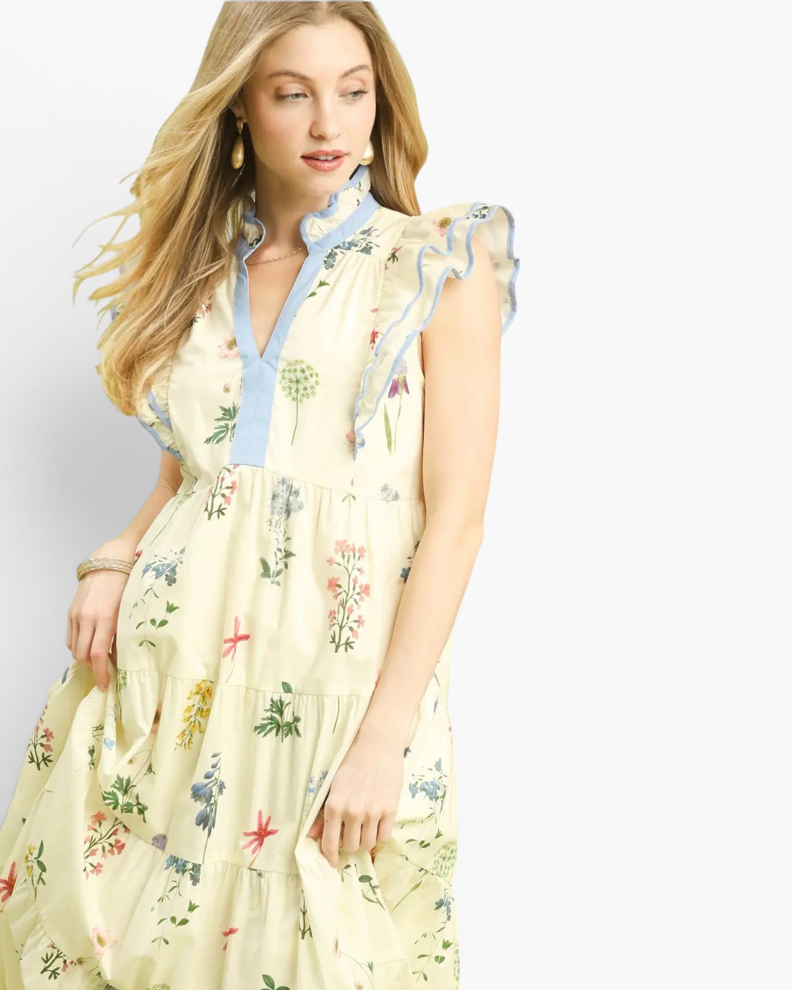 A woman wearing a light cream tiered maxi dress with a blue ruffled collar and a multicolor floral and botanical print, set against a plain white studio background.