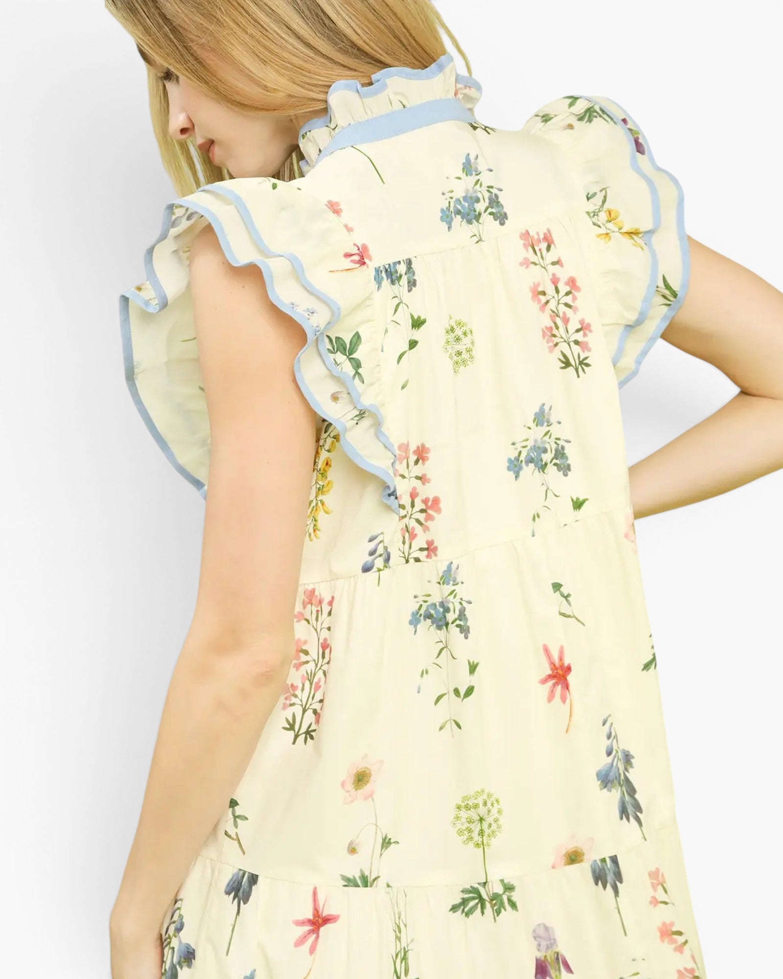 A woman wearing a cream-colored tiered dress with a colorful floral print and light blue ruffled trim, shown from a side-back angle against a plain white background.