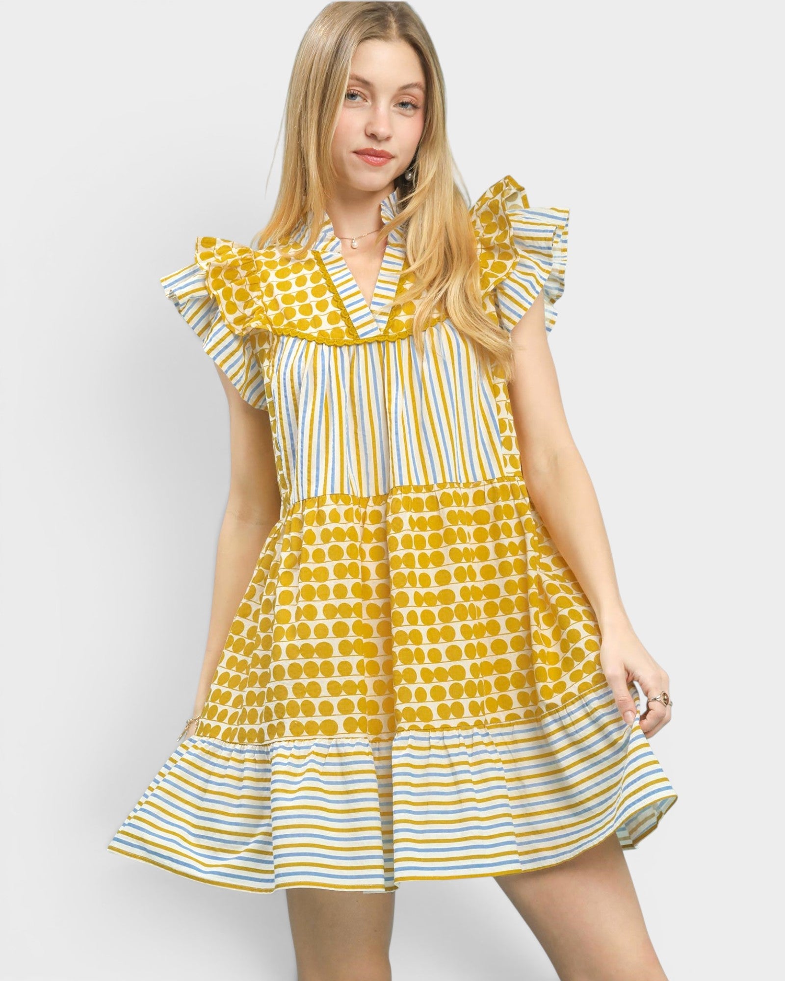 A young woman modeling a yellow and white patterned dress with ruffled sleeves and tiered skirt, photographed against a light gray studio background.
