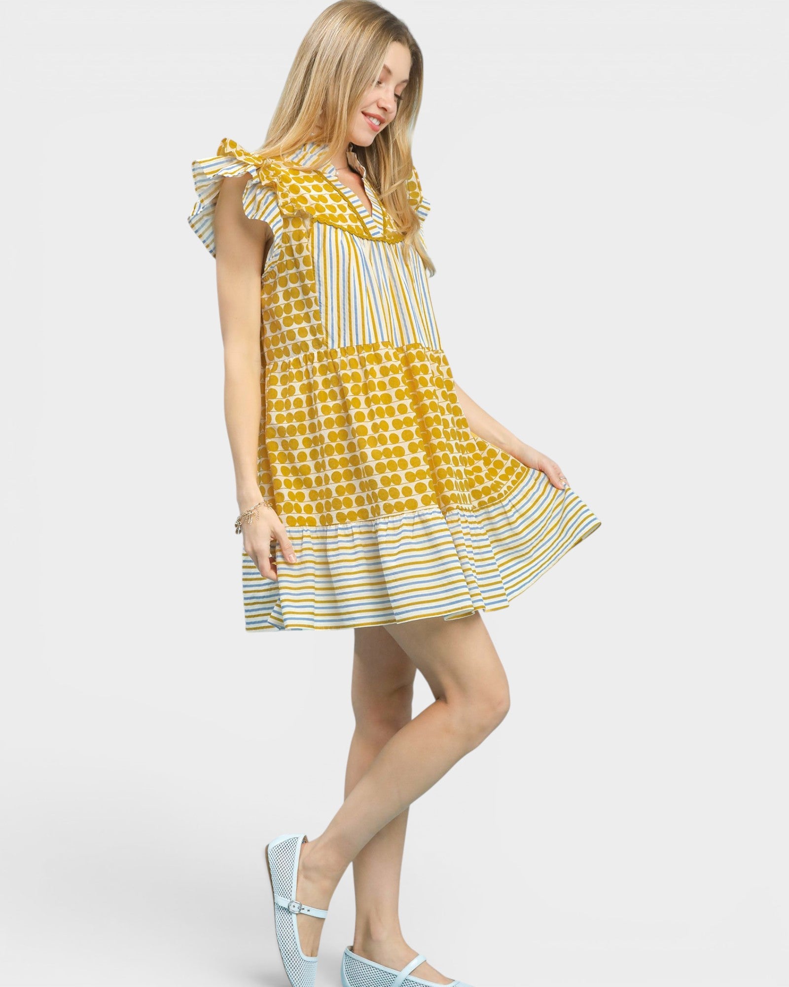 A woman modeling a tiered A-line dress with a yellow and white patterned bodice and striped sleeves and skirt, paired with light blue Mary Jane shoes, against a light gray studio background.