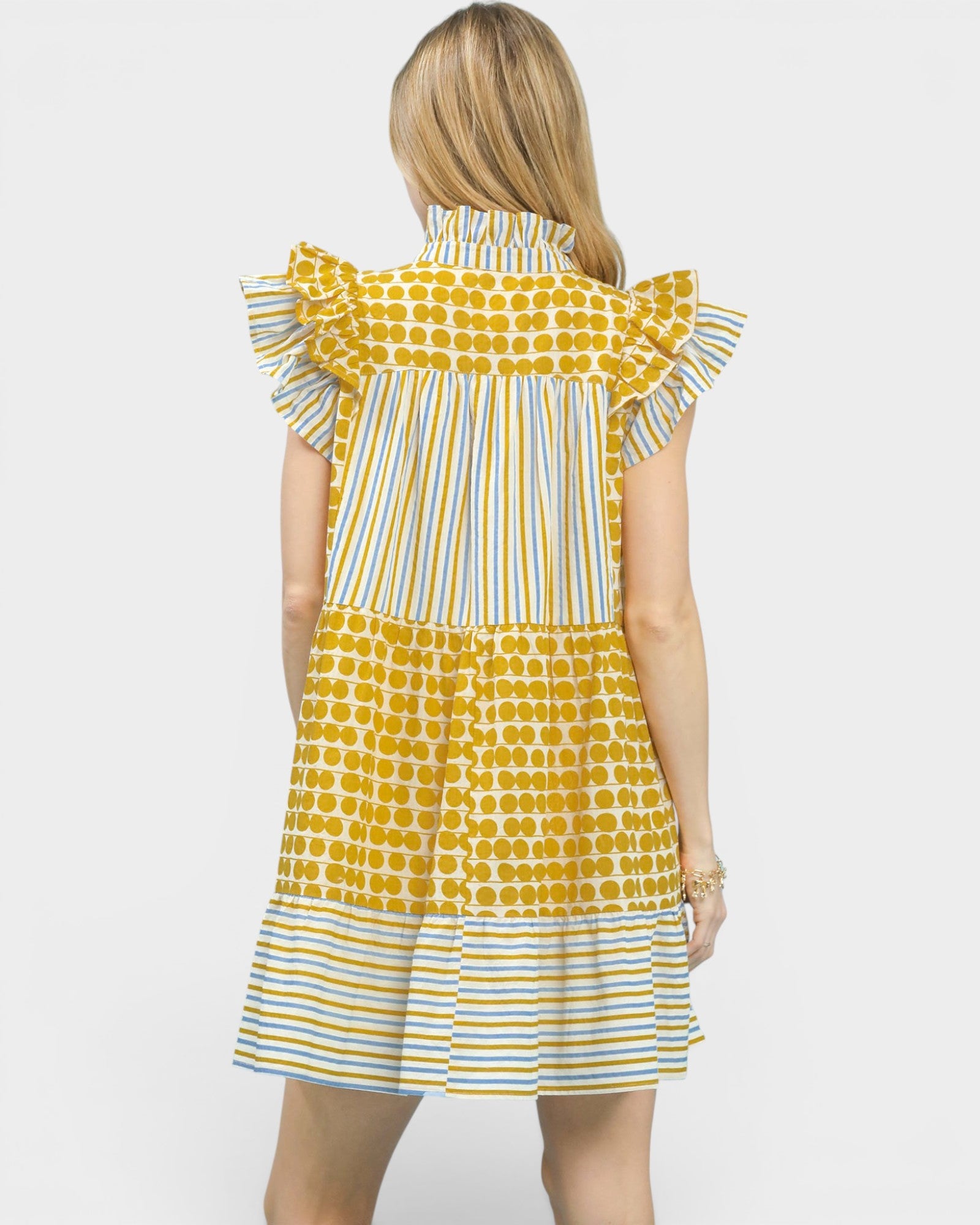 A woman shown from the back wearing a tiered dress with yellow and white striped and polka-dot patterns, set against a plain light gray background.
