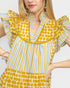 A close-up of a woman wearing a yellow, white, and blue striped dress with ruffled details and a yellow polka-dot pattern on the skirt, set against a plain light gray background.