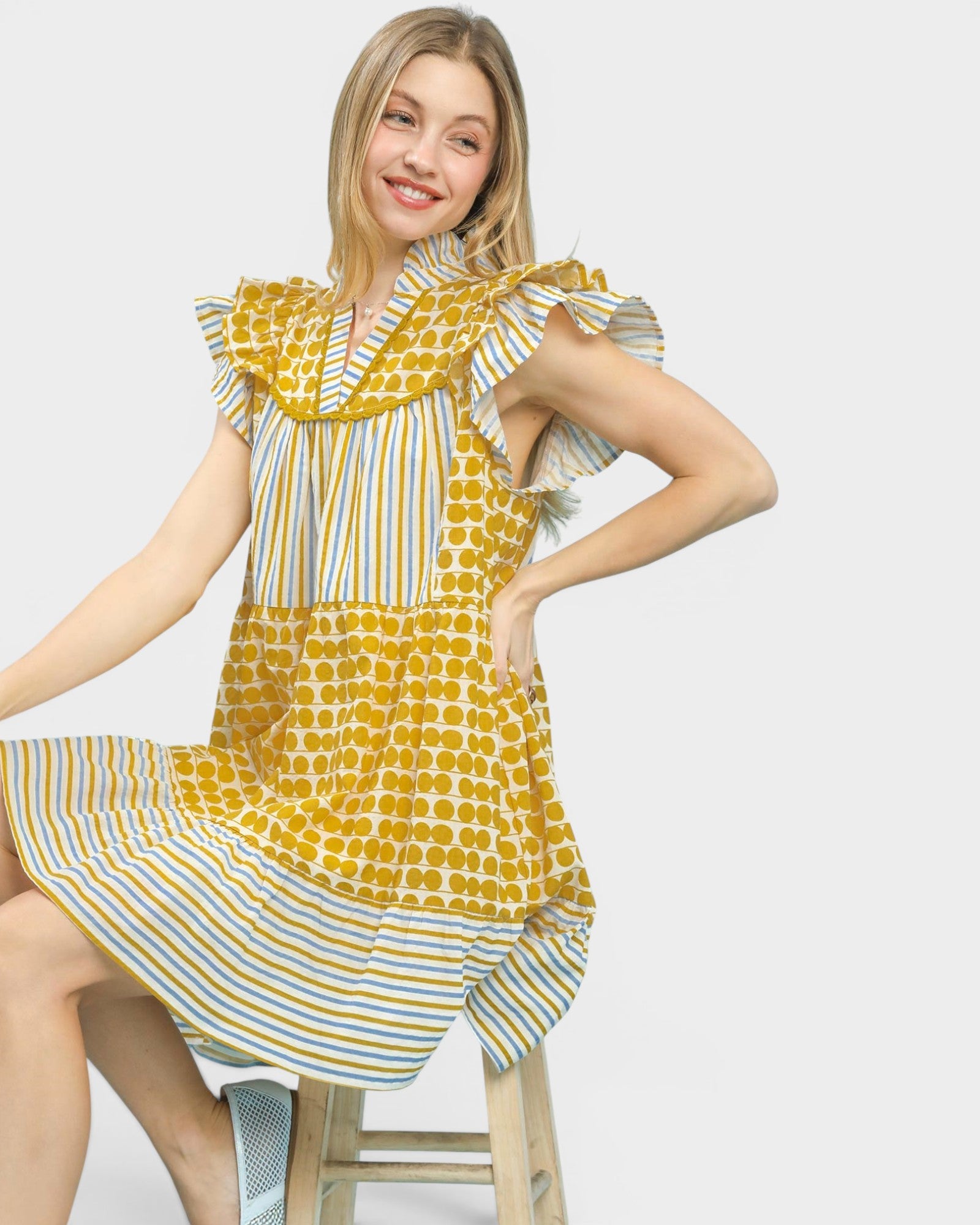 A woman wearing a yellow and white patterned dress with ruffled sleeves, sitting on a wooden stool against a light gray background.