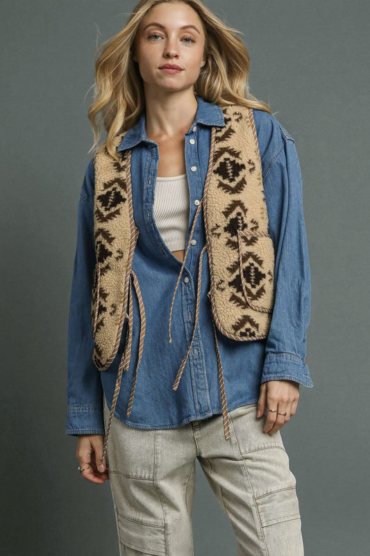 Woman wearing a blue denim shirt, beige patterned vest, and beige pants against a gray background