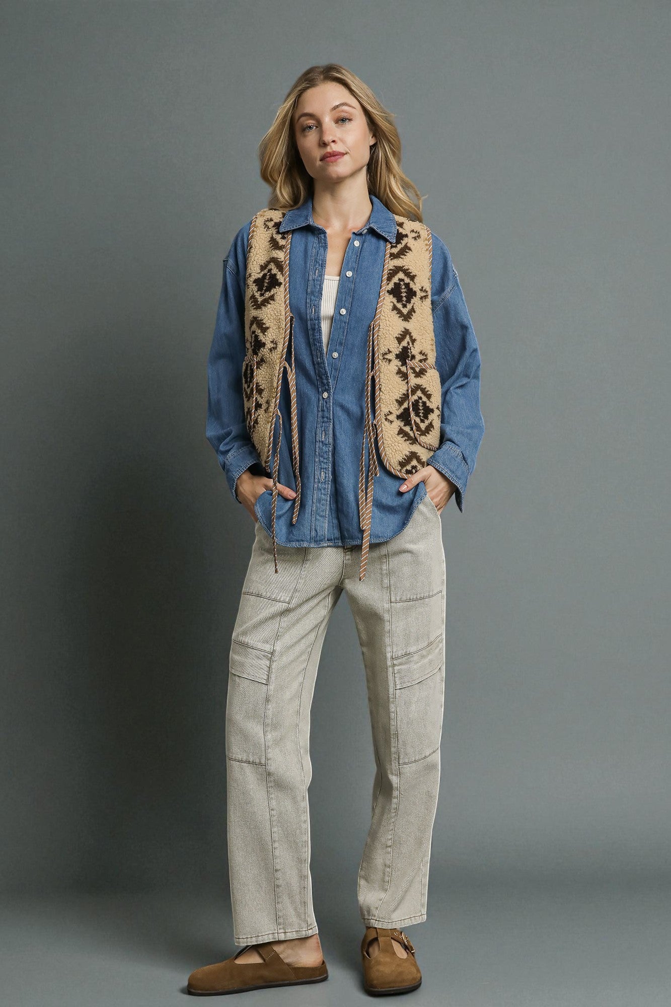 Woman wearing a blue denim shirt, patterned vest, and beige pants against a gray background