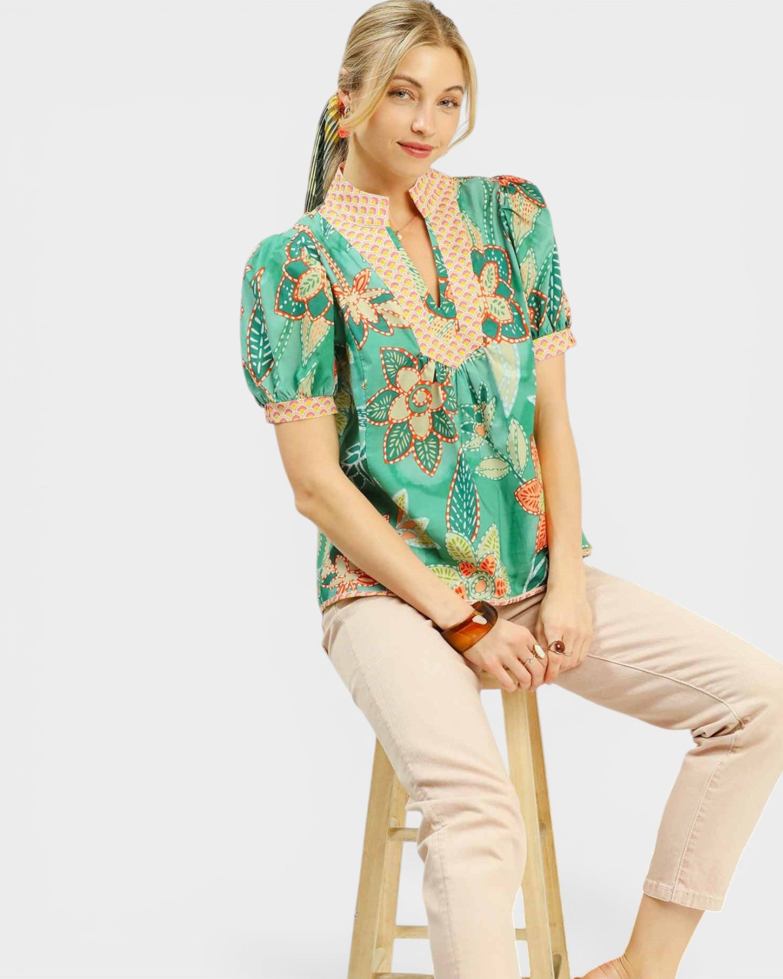 Woman wearing a patterned top and beige pants sitting on a stool against a white background