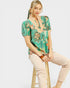 Woman wearing a patterned top and beige pants sitting on a stool against a white background