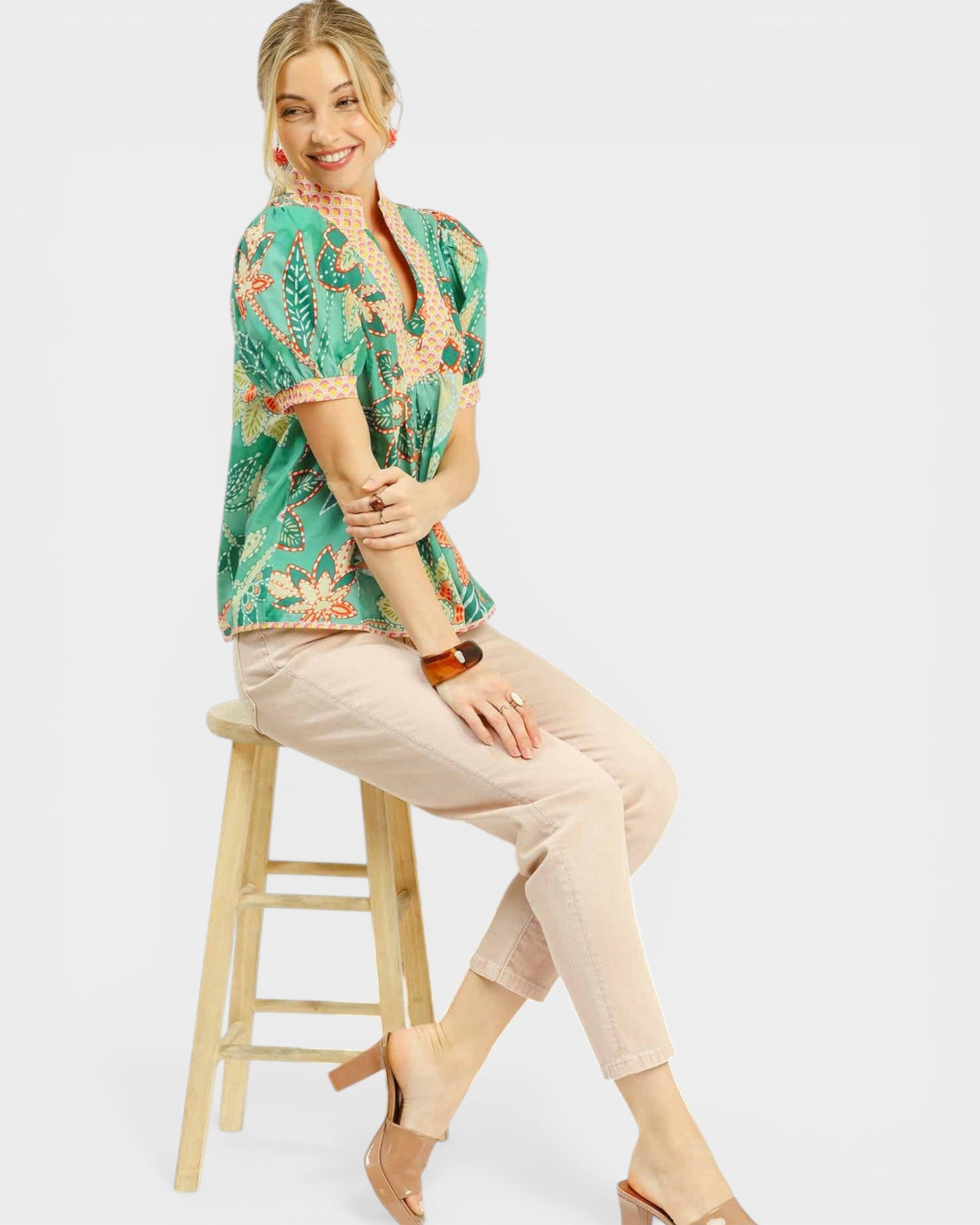 Woman sitting on a wooden stool wearing a floral blouse and beige pants against a white background