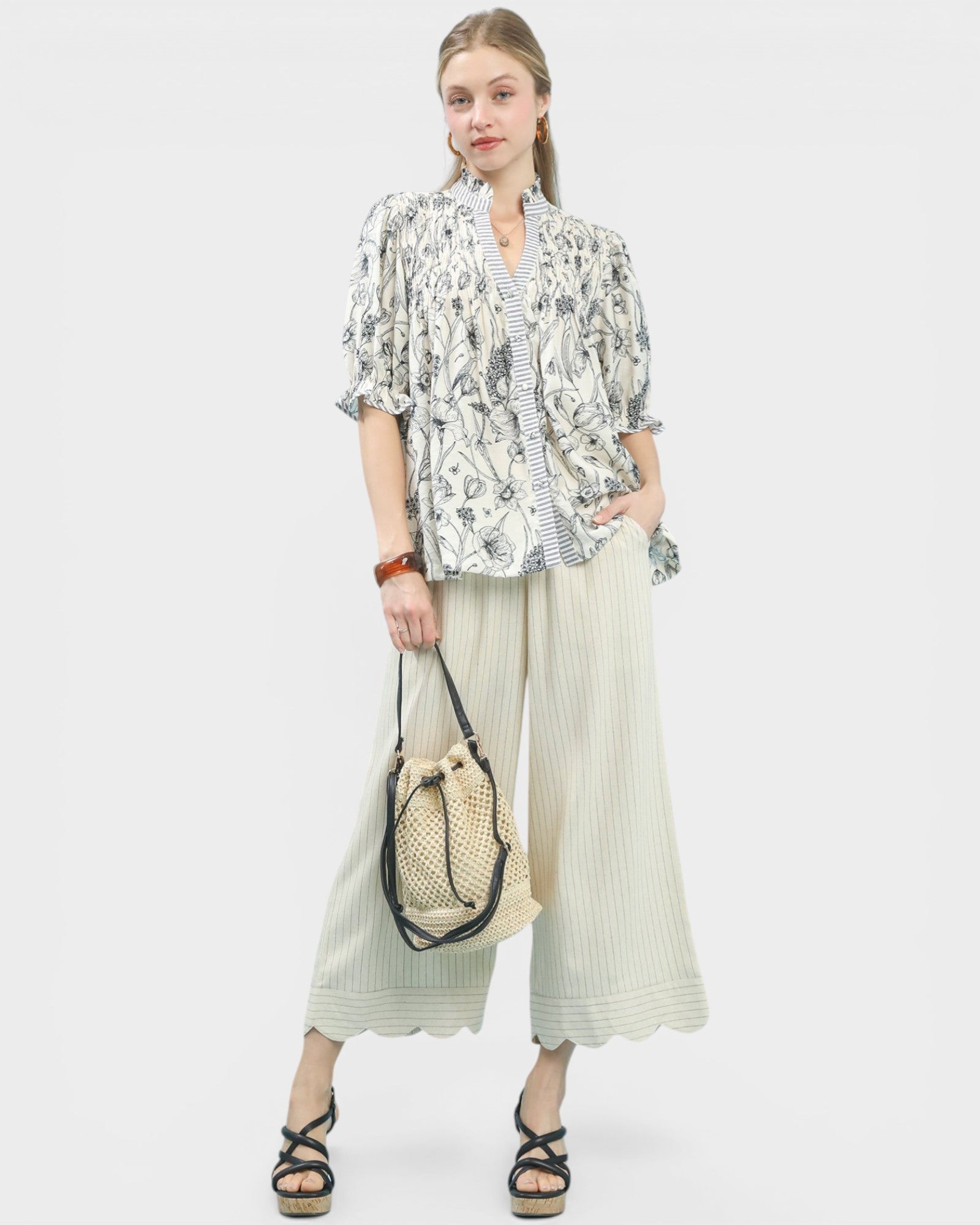 Woman wearing a floral blouse and wide-leg pants holding a woven bag on a white background