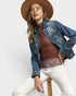 Woman wearing a denim jacket, brown top, and white pants on a white background