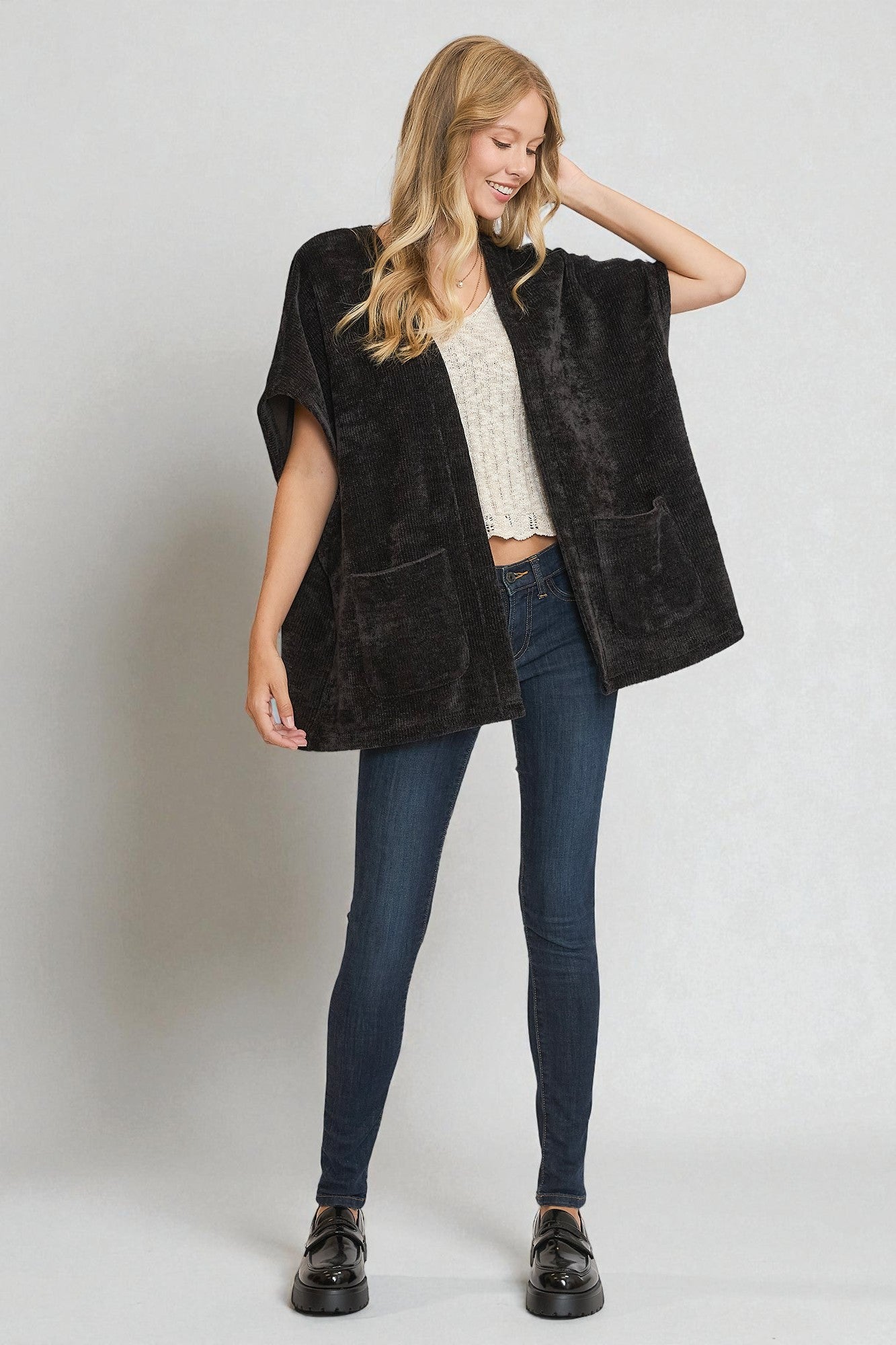 Woman wearing a black chenille vest over a white top and blue jeans on a plain background