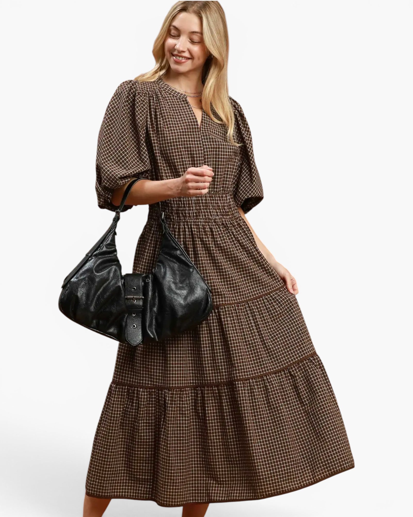 A woman modeling a brown and black checkered tiered midi dress with puff sleeves, carrying a black leather shoulder bag against a light gray studio background.