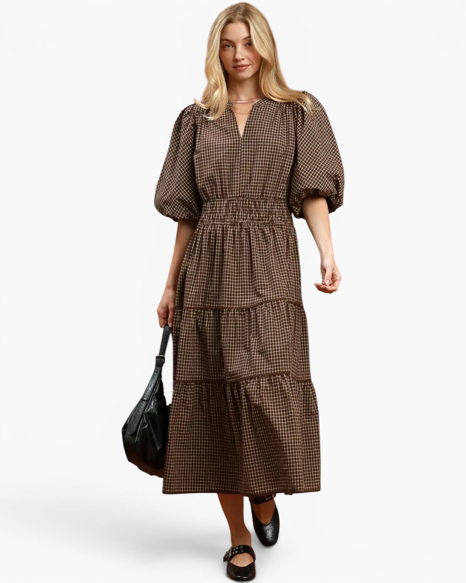 A woman modeling a brown and black gingham midi dress with puff sleeves, a V-neckline, and a tiered skirt, holding a black handbag against a light gray background.