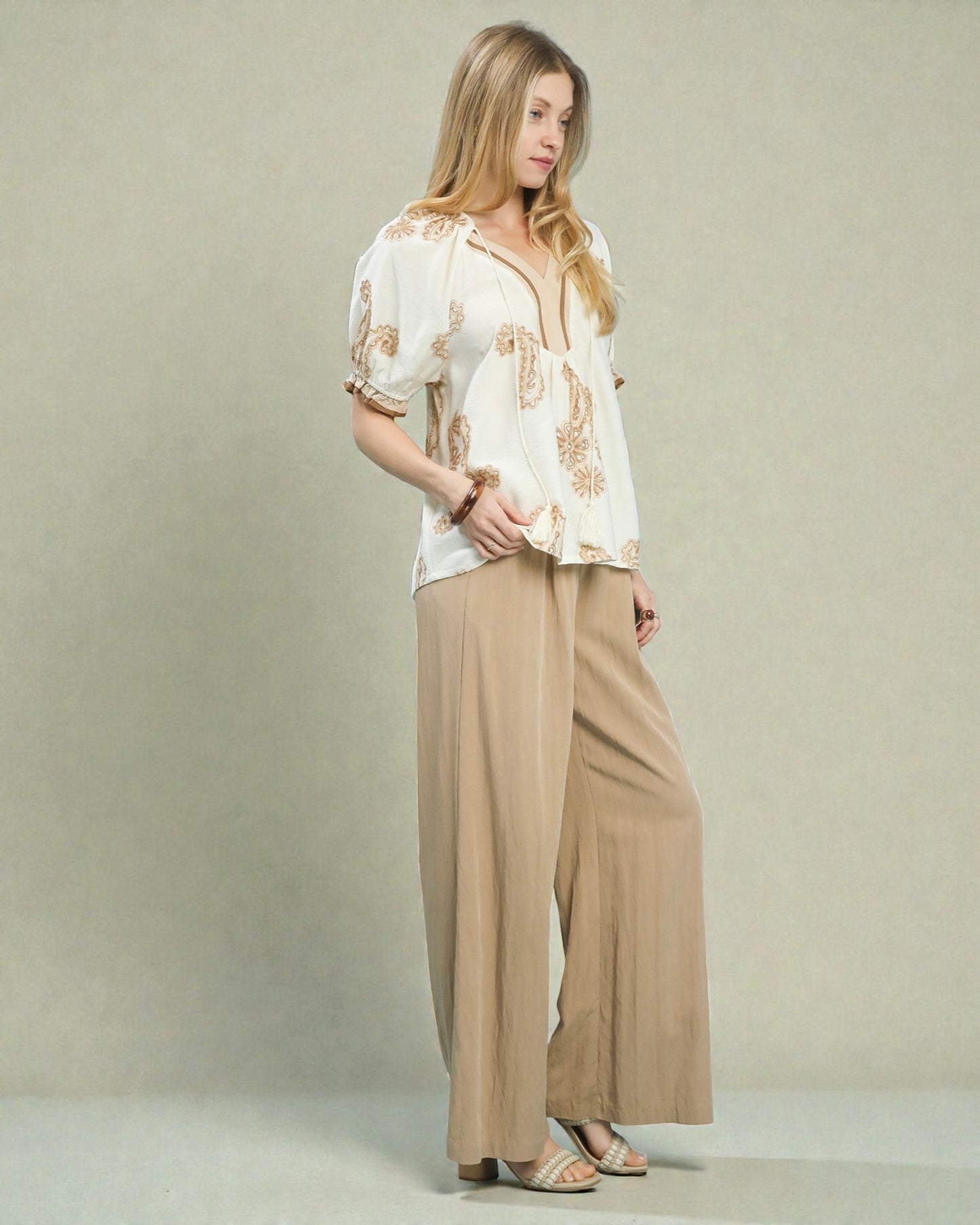 Woman wearing a white blouse with gold patterns and beige wide-leg pants against a plain background