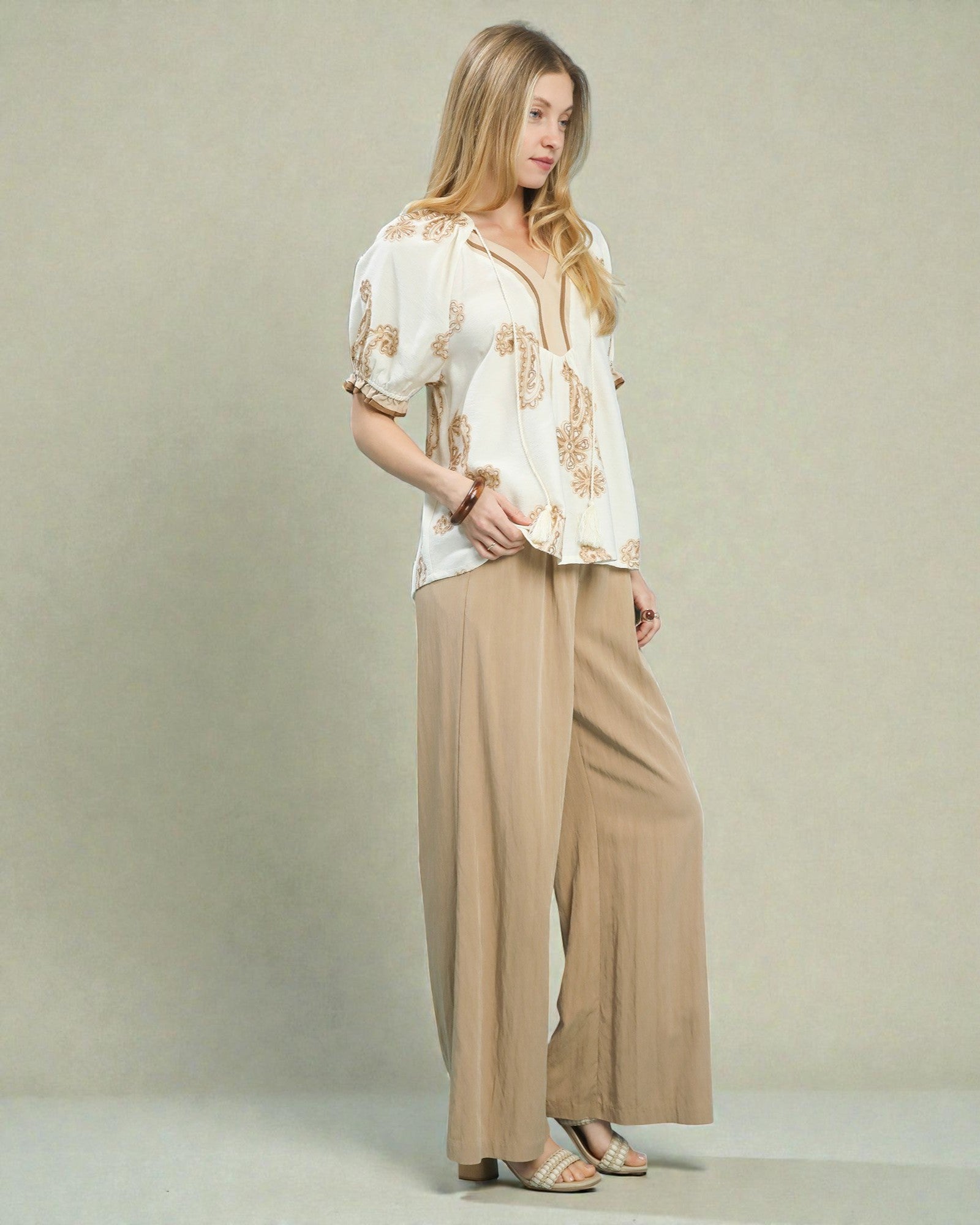Woman wearing a white blouse with gold patterns and beige wide-leg pants against a plain background