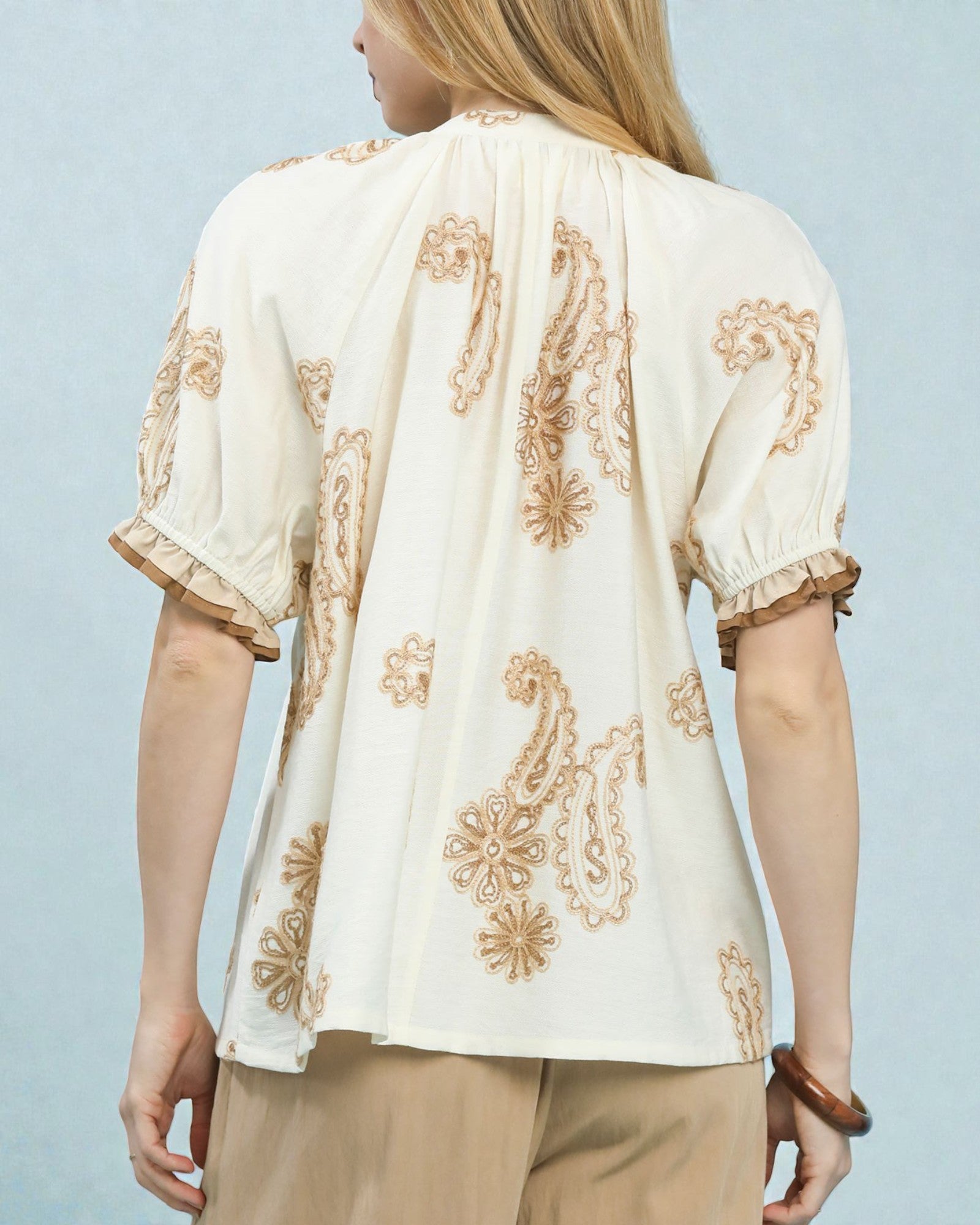 Woman wearing a cream blouse with gold embroidery against a light blue background