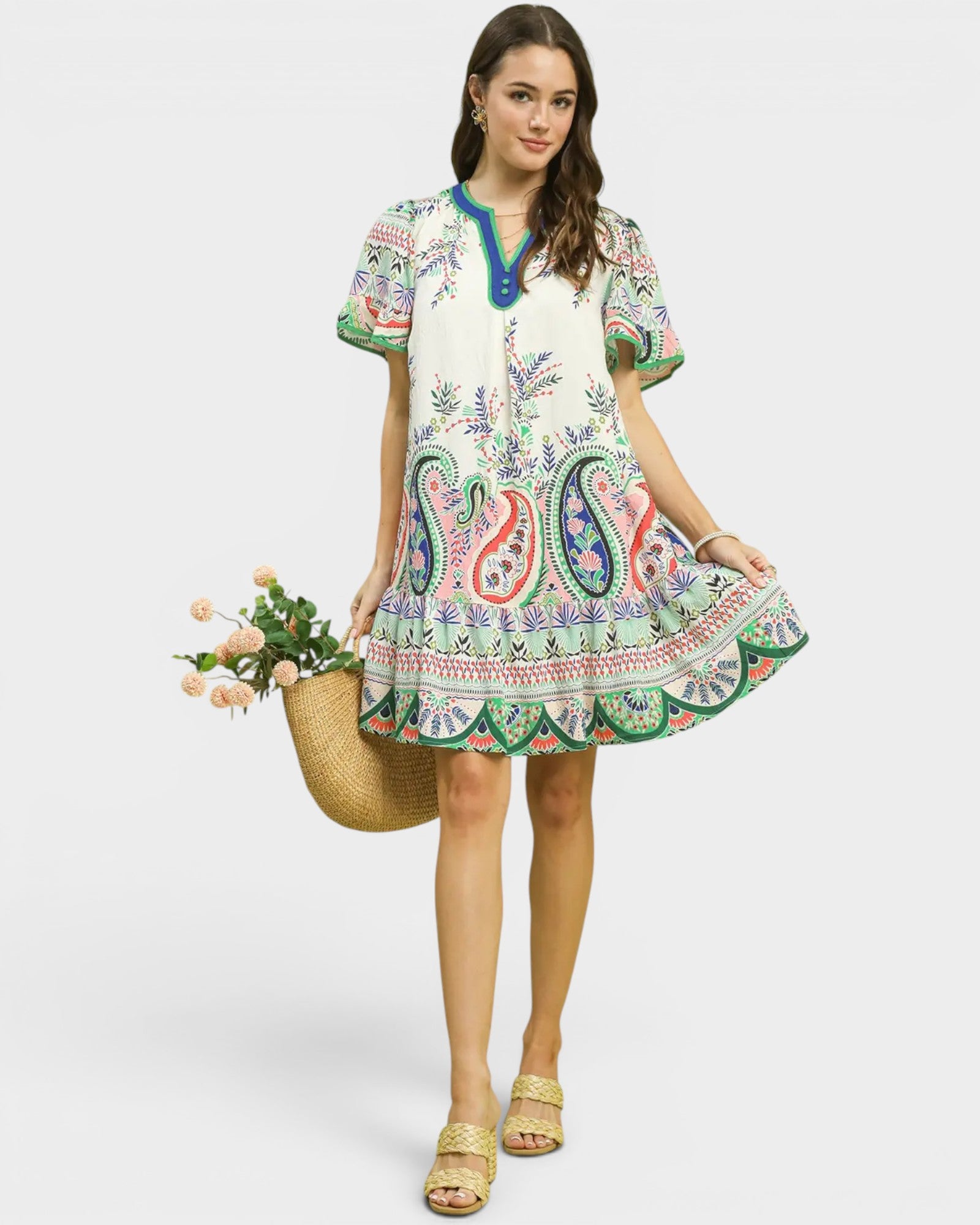Woman wearing a colorful paisley dress holding a woven basket with flowers on a white background