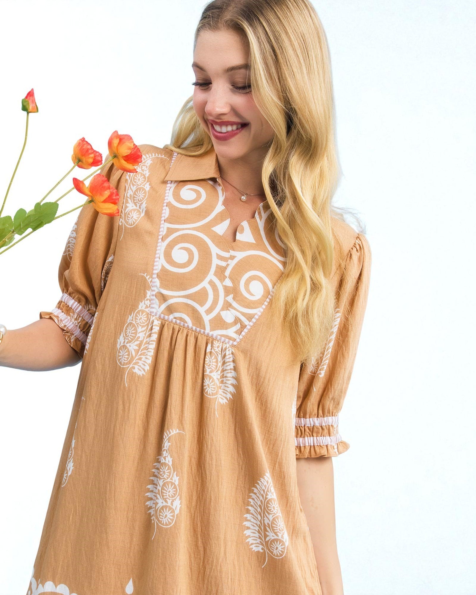 Woman wearing a tan dress with white embroidery, holding flowers against a white background