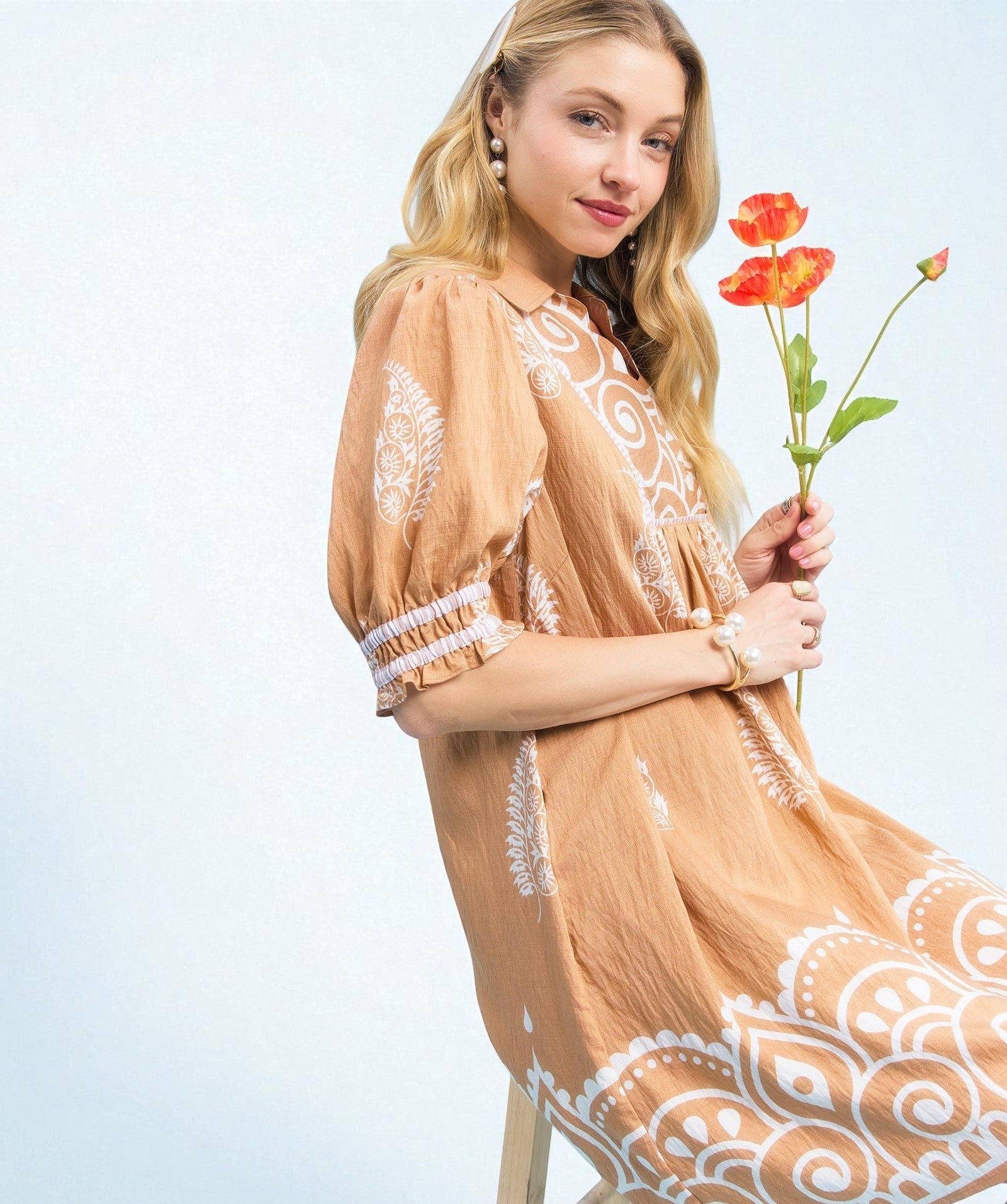 Woman in a beige dress with white lace details holding red flowers against a light blue background