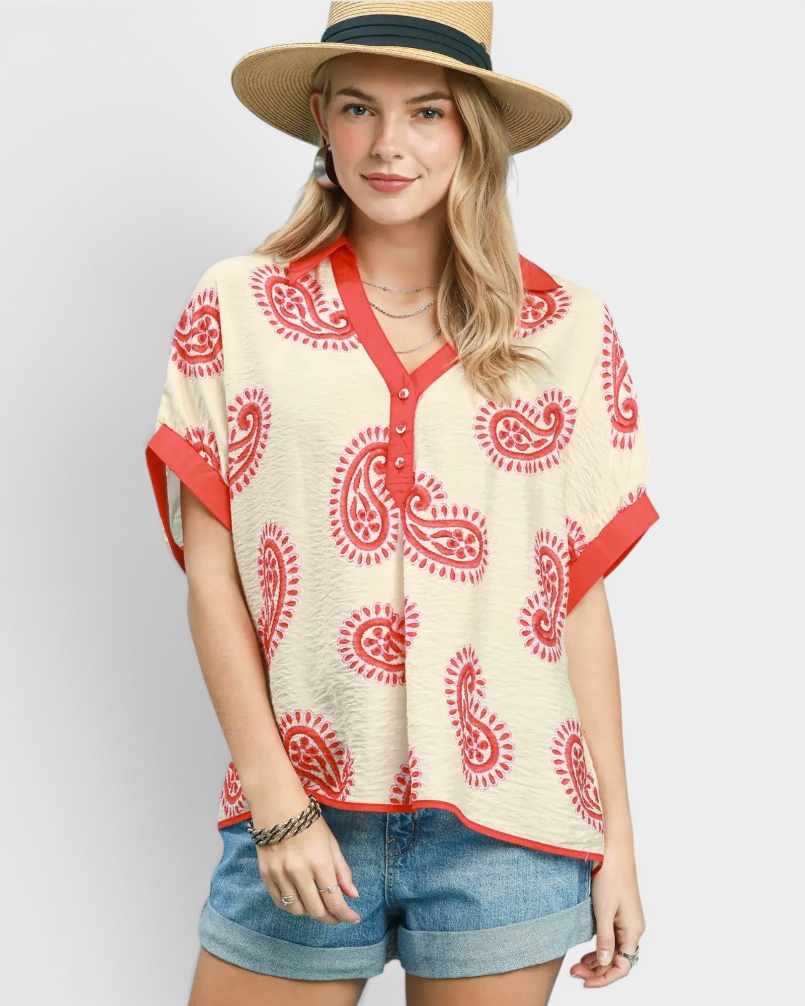 Woman wearing a patterned top with red designs, denim shorts, and a straw hat on a light gray background