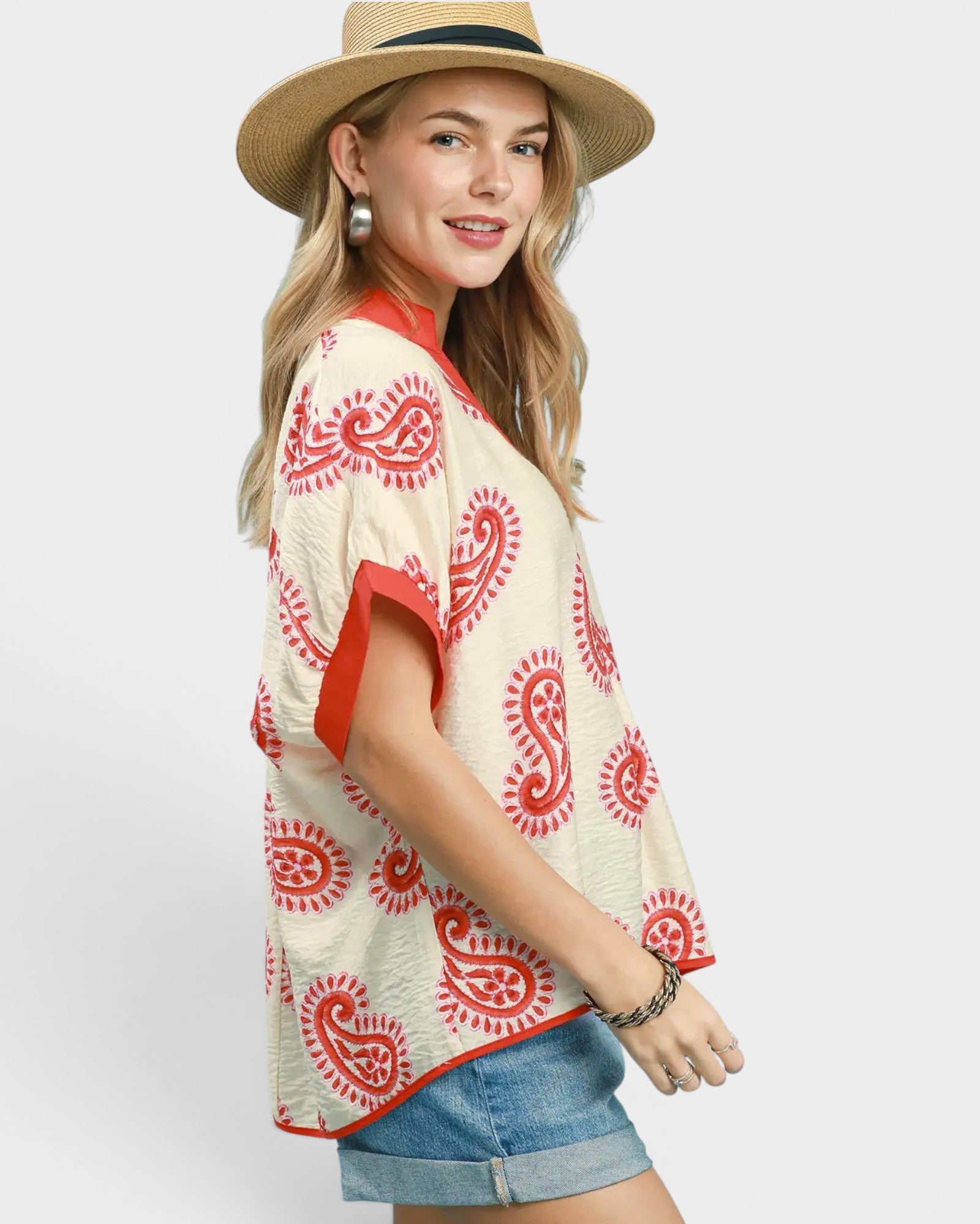 Woman wearing a cream and red patterned top with denim shorts and a straw hat on a white background