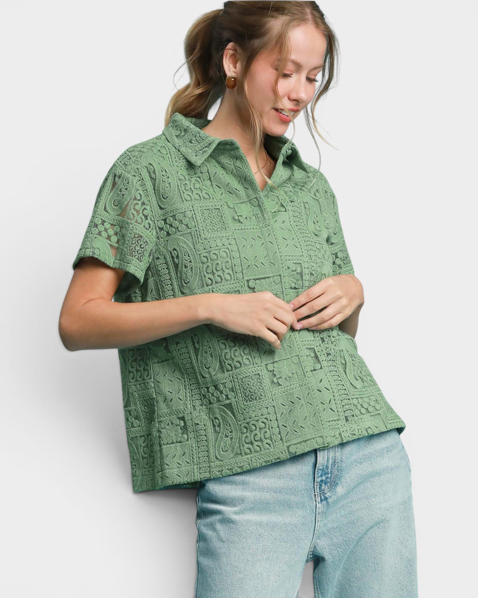 A woman wearing a sage green short-sleeve button-up shirt with an intricate paisley and floral crochet pattern, paired with light blue jeans, against a plain light gray background.