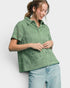 A woman wearing a sage green short-sleeve button-up shirt with an intricate paisley and floral crochet pattern, paired with light blue jeans, against a plain light gray background.
