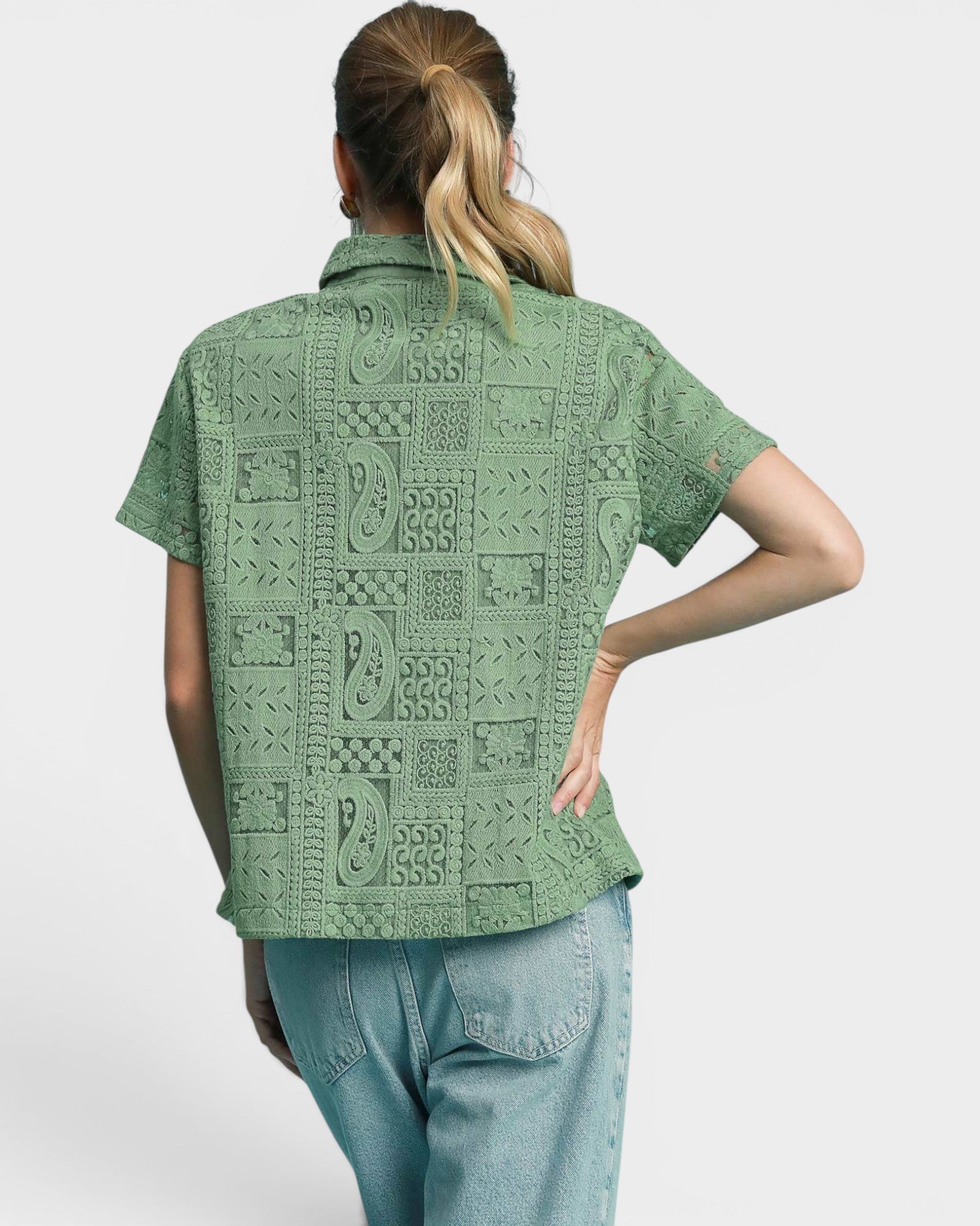 A woman shown from the back wearing a sage green short-sleeve button-up shirt with a textured, raised patchwork pattern, paired with light blue jeans, against a light gray studio background.