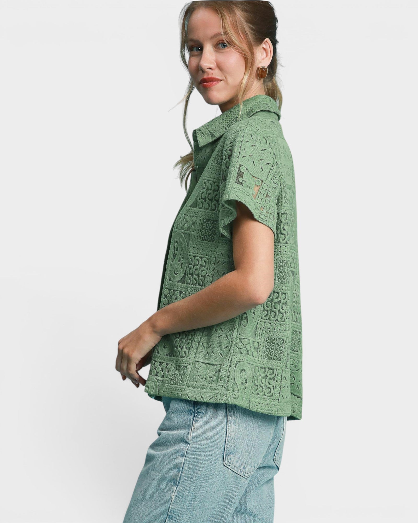 A woman wearing a sage green short-sleeve button-up shirt with a textured, patterned weave, paired with light blue jeans, shown from a side profile against a light gray studio background.
