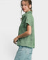 A woman wearing a sage green short-sleeve button-up shirt with a textured, patterned weave, paired with light blue jeans, shown from a side profile against a light gray studio background.