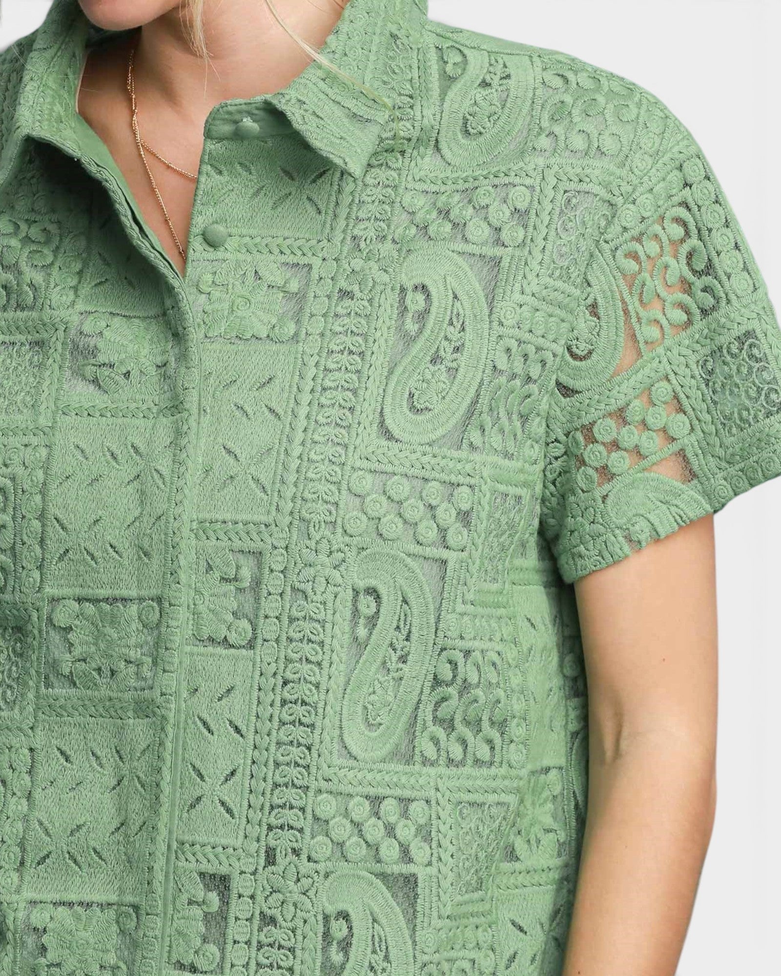 A close-up of a woman wearing a sage green short-sleeve button-up shirt with an intricate lace and crochet paisley pattern.