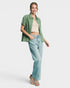 A woman modeling a sage green textured short-sleeve button-up shirt over a beige crop top, paired with light-wash blue jeans and tan sandals, against a solid light gray background.