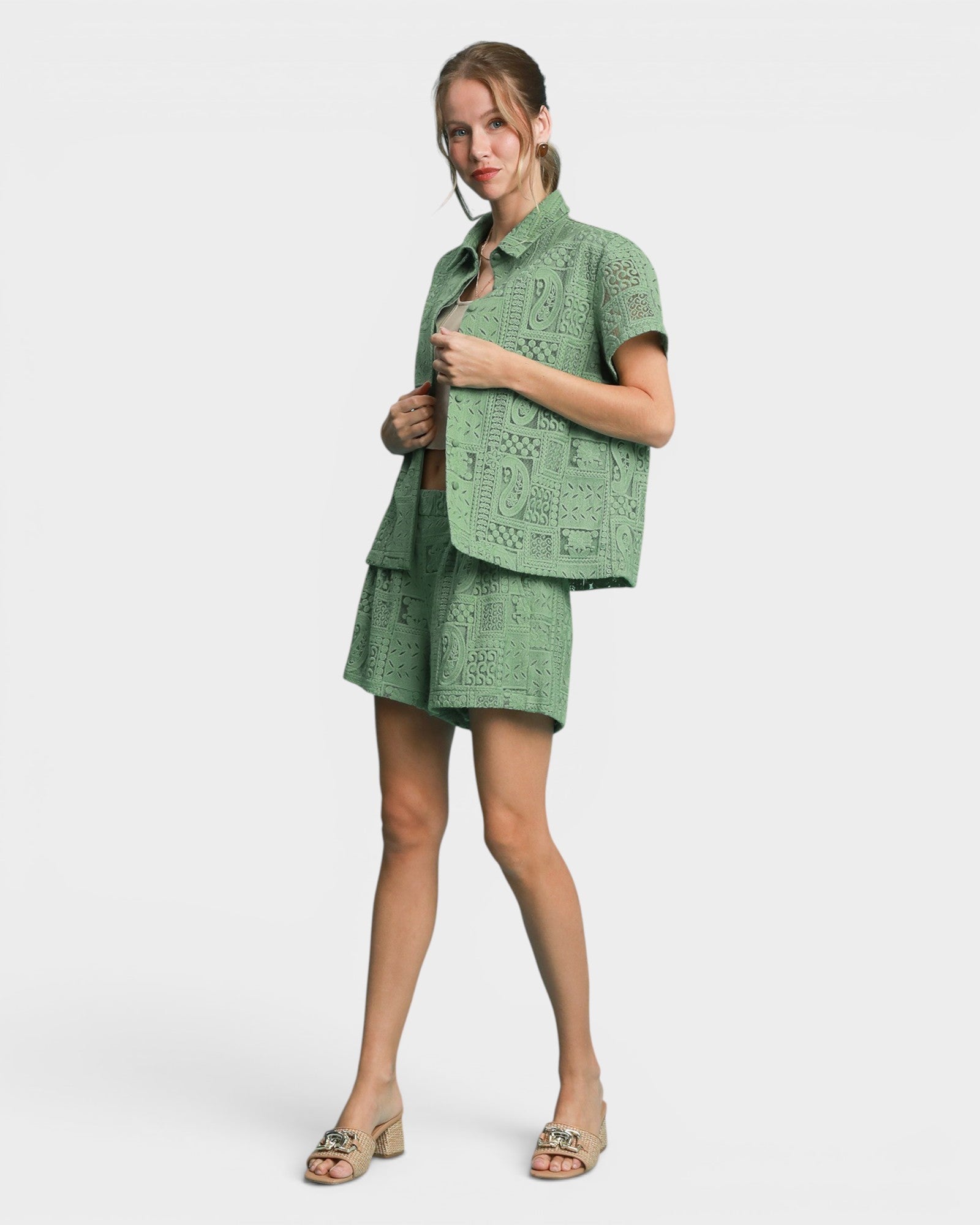 A woman modeling a sage green short-sleeve eyelet knit blazer and matching mini skirt set, paired with beige heeled sandals, against a light gray studio background.