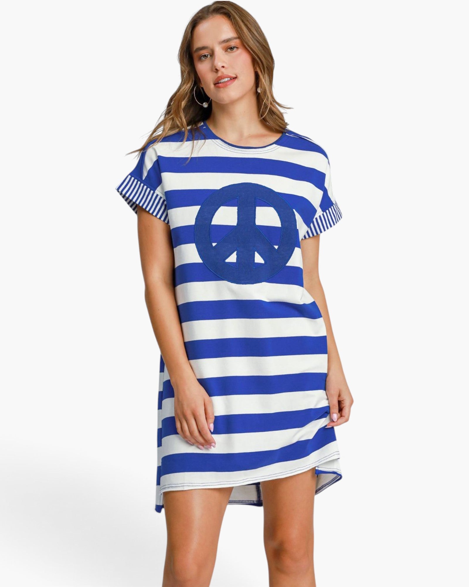 A woman modeling a short-sleeve blue and white striped tunic dress with a large blue peace sign graphic on the front, set against a plain white studio background.
