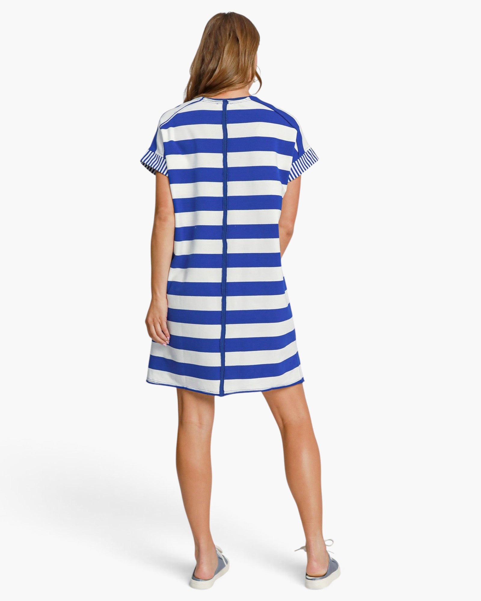 A woman shown from the back wearing a short-sleeve A-line dress with bold horizontal blue and white stripes, paired with gray slip-on shoes, against a light gray studio background.