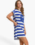 A woman modeling a short-sleeve blue and white horizontally striped dress with a large blue graphic on the front, set against a plain white studio background.