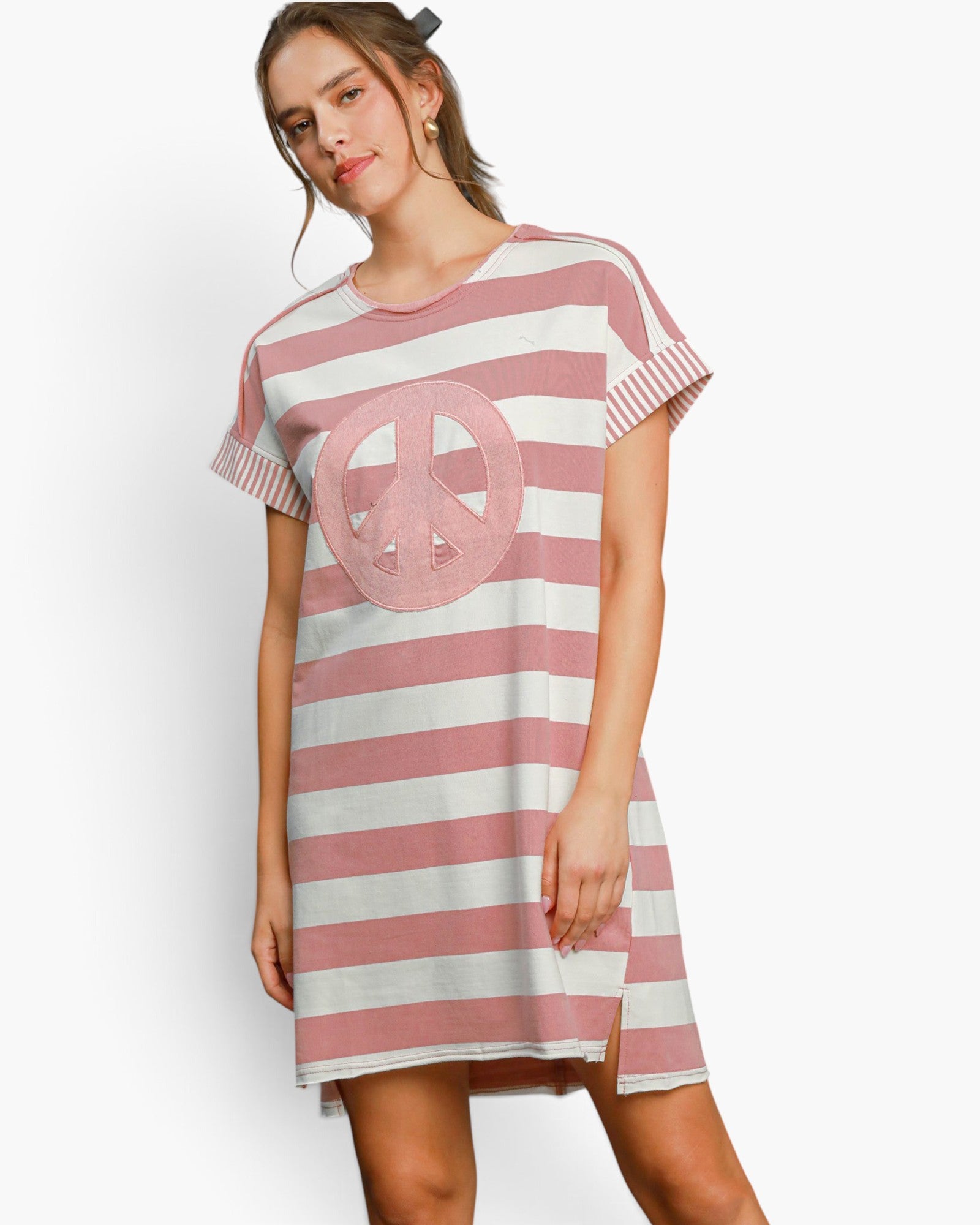A woman modeling a short-sleeve pink and white striped tunic dress with a large pink peace sign applique on the front, set against a plain white studio background.