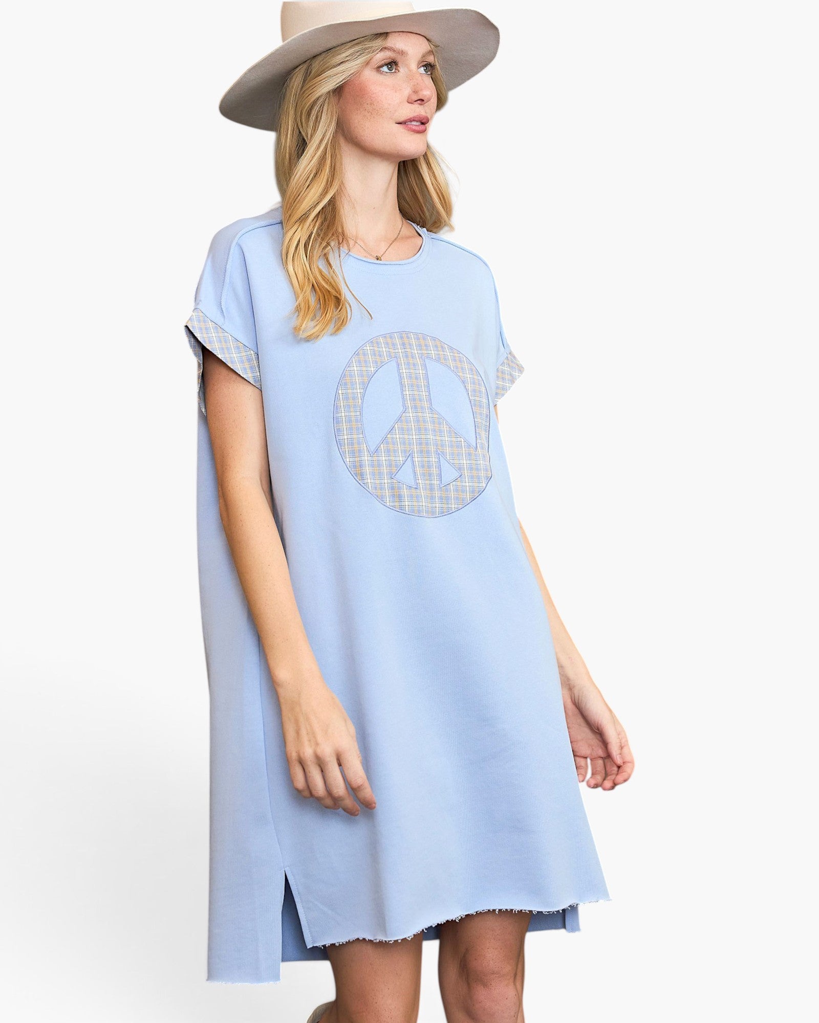 A woman wearing a light blue short-sleeve tunic dress with a large peace sign graphic on the front, styled with a beige wide-brim hat against a plain white studio background.