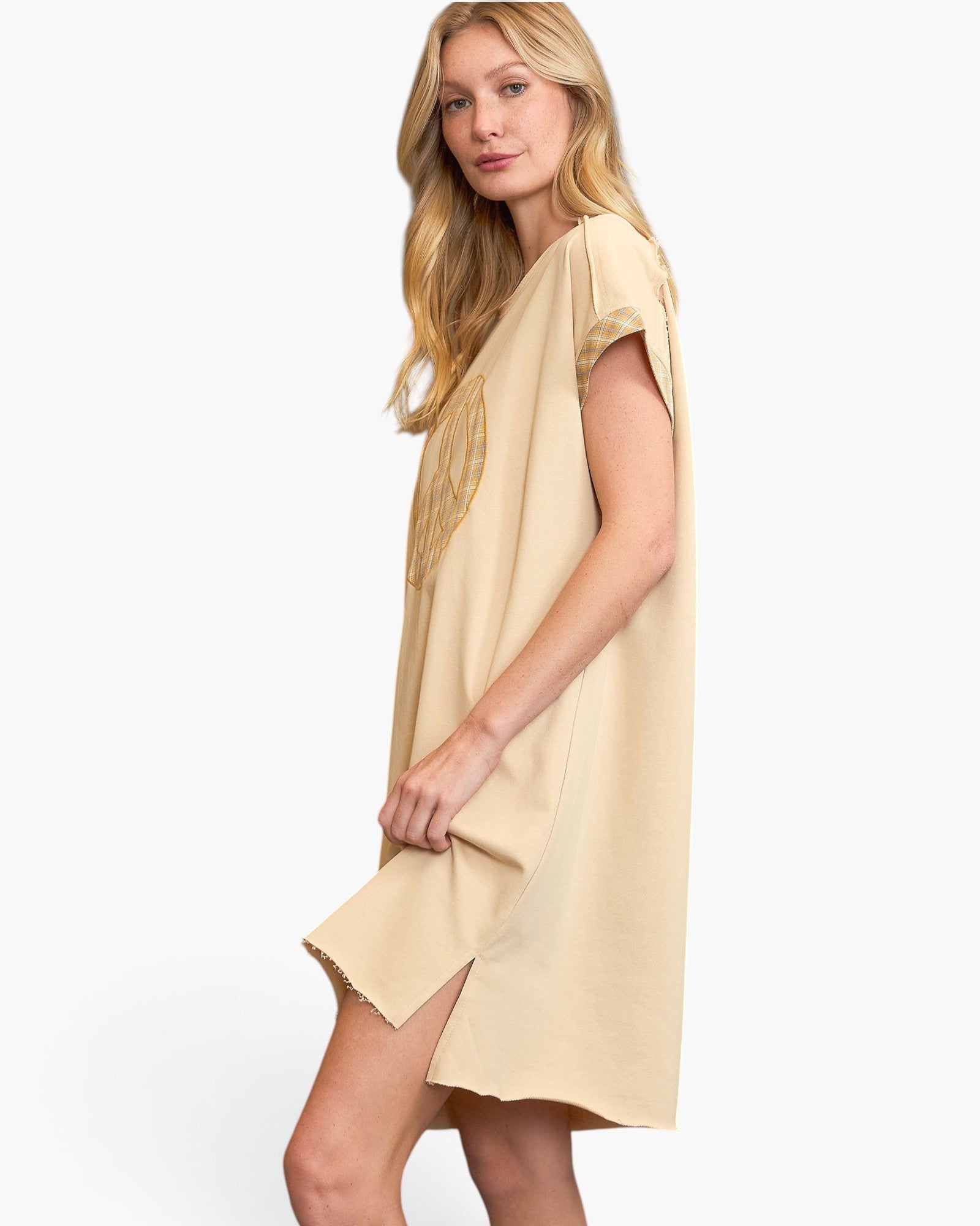 A woman modeling a light beige short-sleeve tunic dress with a relaxed fit and raw-edge hem against a plain white studio background.