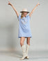Woman wearing a light blue dress with a peace symbol, white cowboy boots, and a beige hat against a gray background.