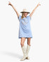 A woman modeling a light blue short-sleeve tunic dress with fringe and a peace sign graphic on the front, styled with a beige wide-brim hat and cream-colored cowboy boots against a light gray studio background.